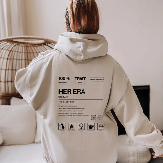 Back view of a young woman wearing Urban Cookie's HER ERA Heavy blend Sand Blueprint Hoodie in a neutral tone living room with natural  lighting.