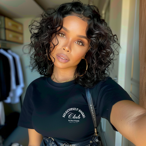 Young trendy woman with short curly hair and big gold hoops wearing an Urban Cookie  Black Selectively Social small print wavy White text tee. Blurry closet background.